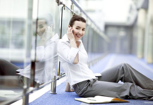 Atractive young woman sitting on the floor in modern office building corridor with mobile phone in her hand