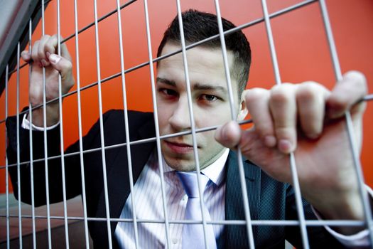 Portrait of young agressive man in business clothes behind bars with red backgroung