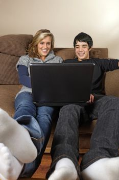 Two happy teenagers, Caucasian female and Asian male, sitting on a couch surfing the internet.