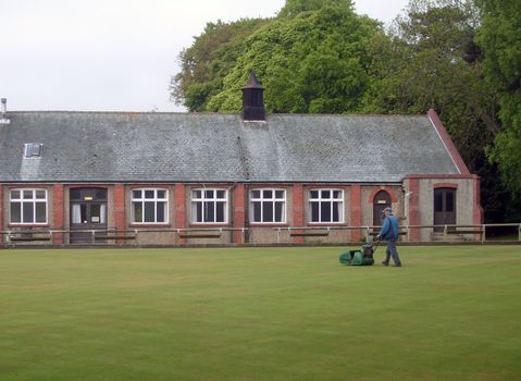 English village bowling green and pavilion on summers day, with groundsman cutting grass.