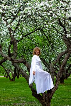 The blonde girl in white dress sittng on apple-tree with white flowers
