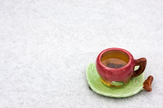 Iin the lower left corner of the frame is a cup of black tea on a snowy background. Saucer shaped leaf.