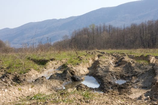 Dirt forest road with big puddles.The road washed out by rain. On the road, traces of large machines. Russian off-road. In the background - mountains and forests without leaves.