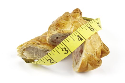 Single golden sausage roll cut in half with pork pie and yellow tape measure on a reflective white background