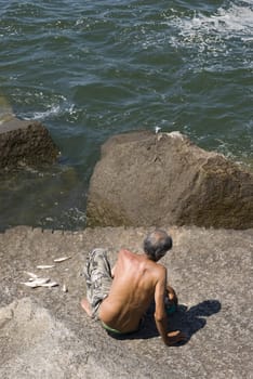 High angle rear shot of an old man fishing on top of rocks, with sea directly below.
