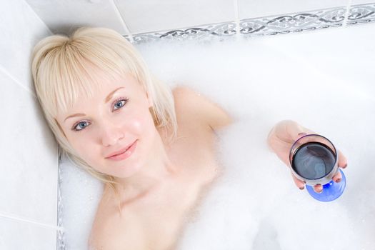 a young woman baths in a bathroom in much foam and drink wine.