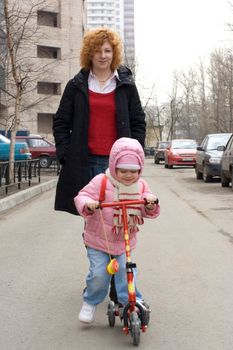Little girl on the scooter and her young mother walking on the street