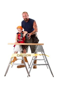 Caucasian dad is teaching cute son with construction helmet how to drill a hole in a wooden plank, isolated.