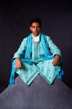 Beautiful authentic Indian hindu man in typical ethnic groom attire sitting in lotus position with legs crossed on top of rock. Bangali male wearing a light blue agua decorated Dhoti with shawl.