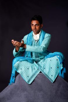 Beautiful authentic Indian hindu man in typical ethnic groom attire sitting in lotus position with legs crossed on top of rock and sticking hands out to receive a gift. Bangali male wearing a light blue agua decorated Dhoti with shawl.