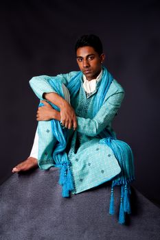 Beautiful authentic Indian hindu man in typical ethnic groom attire sitting relaxed on top of rock leaning on knee. Bangali male wearing a light blue agua decorated Dhoti with shawl.