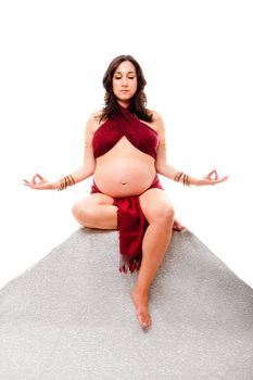 Beautiful pregnant woman with bindi sitting on a stone pyramid with eyes closed dressed in red and meditating, isolated.