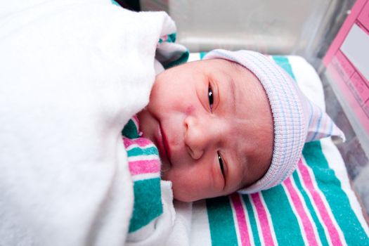 Cute face of a innocent Caucasian newborn baby with open black eyes, wearing a unisex hat, laying on white with green and pink sheets.