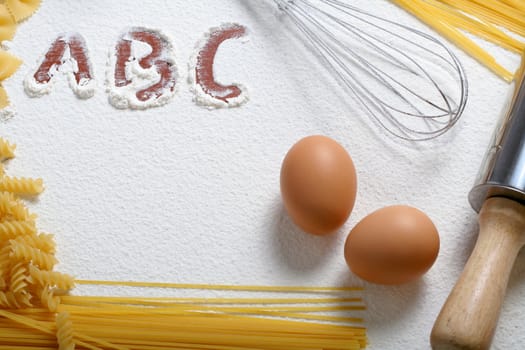 Uncooked macaroni, whisk and eggs on wheat flour, background