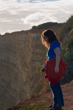A woman with a camera at the edge of a cliff, looking out, with more cliffs in the background.  Near Lagos, Portugal.