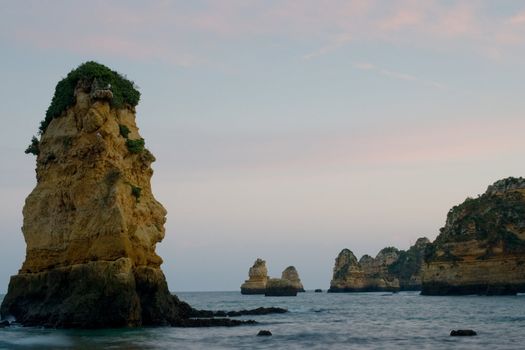 Along the beaches in Lagos, Portugal, a number of sheer rocky outcrops, as the sun sets casting pinks and blues in the sky.
