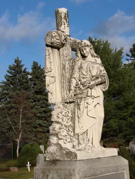 White marble statue of Mary in Catholic Cemetery. Statue has moss and lichen on it. Fall day with bright blue sky and clouds.