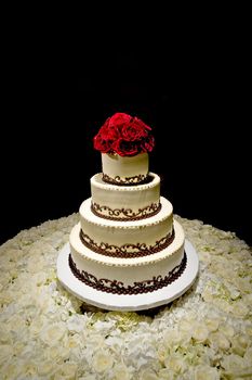 Image of a traditional four tiered wedding cake with red roses on top sitting on a bed of white roses