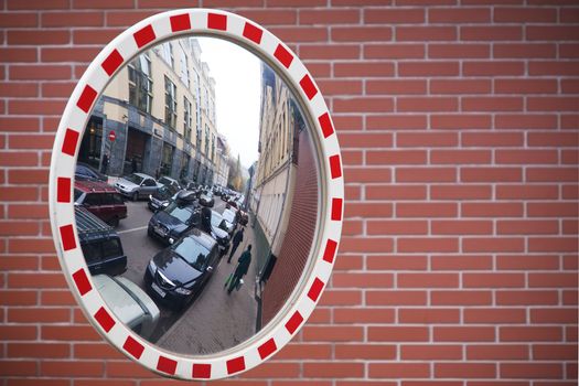 The street with cars and people is reflected in a mirror on a brick wall