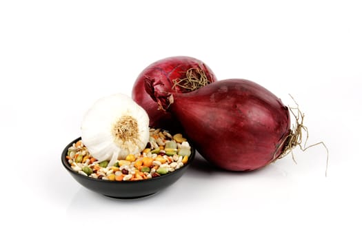 Two raw unpeeled red onions with soup pulses in a small black dish and garlic on a reflective white backgrounds