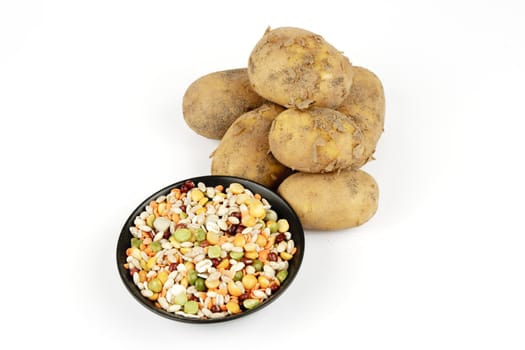 Small pile of brown unpeeled potatoes with a small black dish of soup pulses on a reflective white background