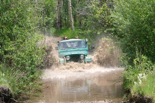 Young rider holds his arms above the roof of a popular vintage four-wheel-drive vehicle while fording a stream across a mountain back-road trail.