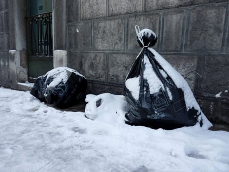 Garbage bags covered by snow in the street next to a building