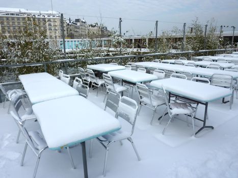 Tables and chairs from a terrace under the snow with city buildings in the background