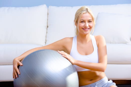 Young woman in sportswear holds an exercise ball while sitting in a living room.  Horizontal shot.