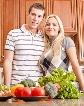 Low angle view of young couple smiling into the camera from behind a kitchen counter. The counter is holding a selection of fresh vegetables. Vertical shot.
