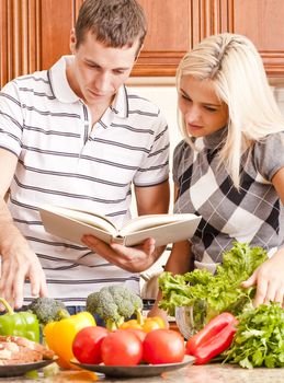 Young couple follows a recipe book. The kitchen counter is full of fresh vegetables. Vertical shot.