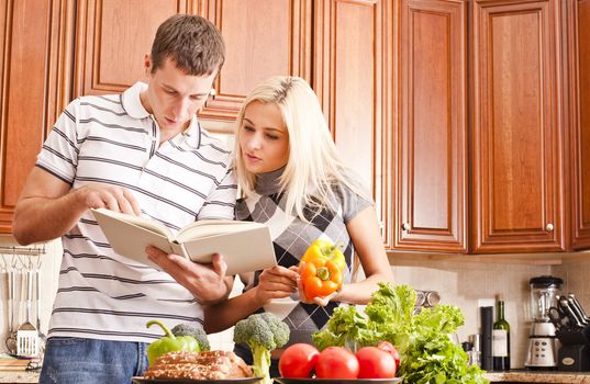 Young couple in the kitchen read an open recipe book. The counter is full of fresh vegetables. Horizontal shot.