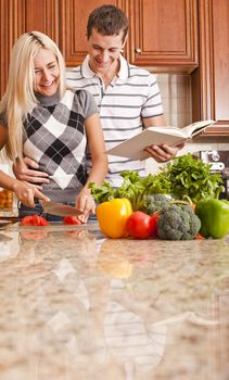 Young woman cutting tomato standing next to man with cookbook. Vertical shot.