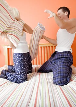 Young couple facing each other, kneeling on bed having a pillow fight. Vertical shot.
