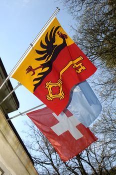 Three flags. The flag of the Swiss canton of Geneva in front, the swiss Conferderation flag behind and, just visible between them, the pale blue flag of the United nations