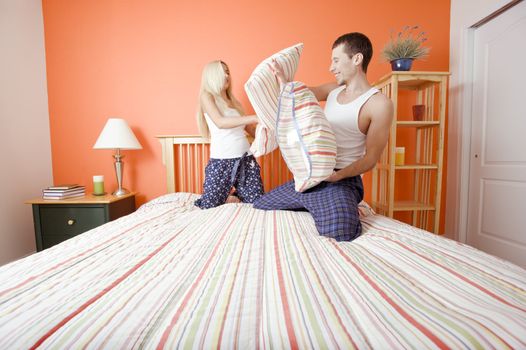 Young couple facing each other, kneeling on bed having a pillow fight. Horizontal shot.