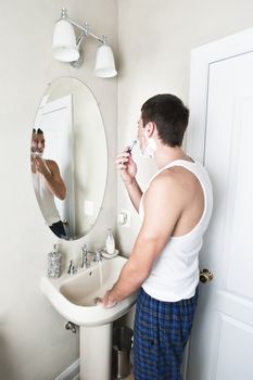 Young man in bathroom looking in the mirror and shaving. Vertical shot.