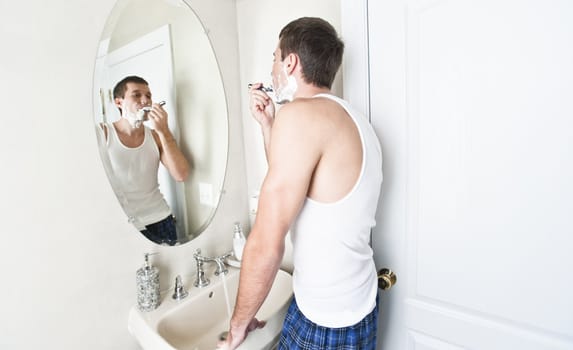 Young man in bathroom looking in the mirror and shaving. Horizontal shot.