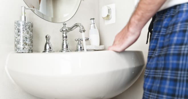 White porcelain sink with water running from the faucet and waist of man in shot. Horizontal shot.
