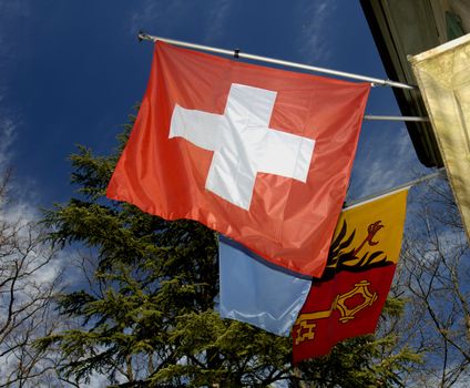 The flag of the Swiss Confederation with the flag of the canton of Geneva behind, against a blue sky laced with fine clouds.
