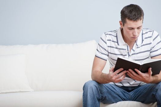 Young man in striped shirt and blue jeans sitting on white sofa reading a book. Horizontal shot.