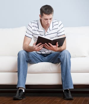 Young man in striped shirt and blue jeans sitting on white sofa reading a book. Vertical shot.