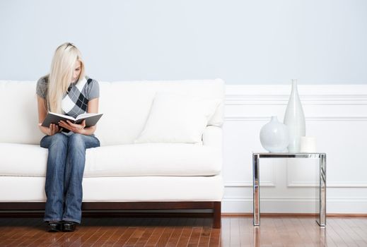 Young woman in checkered shirt and blue jeans sitting on white sofa reading a book. Horizontal shot.