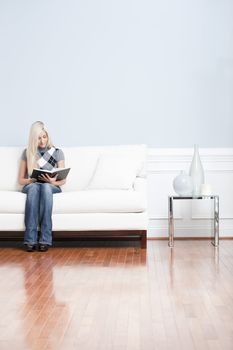 Young woman wears a checkered top and blue jeans while sitting on white sofa.  She is reading a book. Vertical shot.