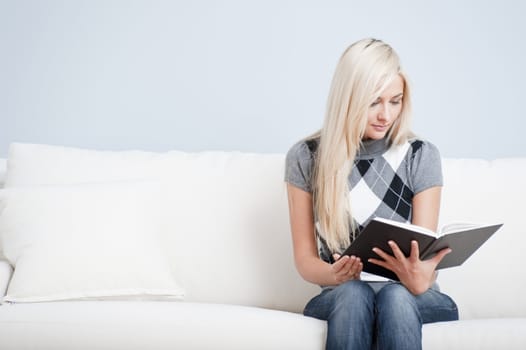 Young woman wears a checkered top and blue jeans while sitting on white sofa.  She is reading a book. Horizontal shot.