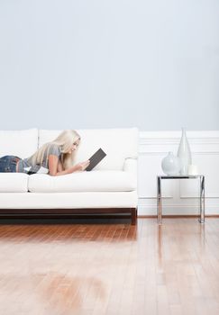 Young woman lies on a white sofa in a checkered top and blue jeans while reading a book. Vertical shot.
