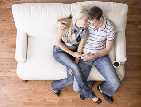 Young couple sit on a cream colored love seat. The man has his arm around the woman. Horizontal shot.