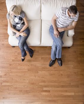 Man and a woman sit distantly on the ends of a cream colored love seat. Their heads are turned away from each other. Vertical shot.