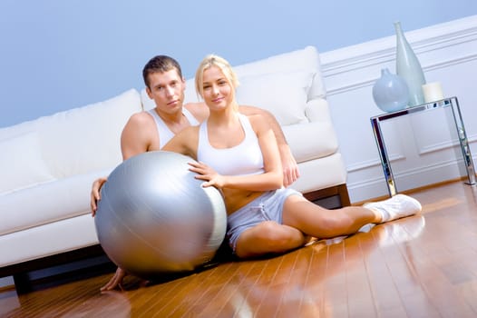 Young couple sit on the wood floor, smiling and holding a silver exercise ball. Horizontal shot.