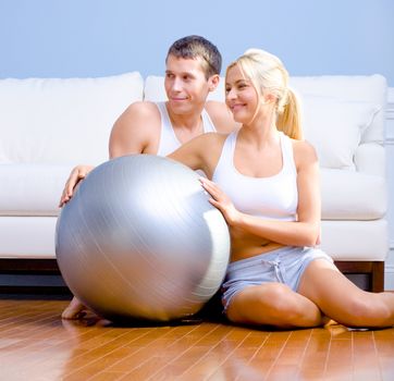 Young couple sit on the wood floor, smiling off to the side while holding a silver exercise ball. Horizontal shot.